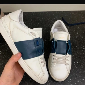 Valentino Garavani vita sneakers med blå detalj - Säljer ett par vita sneakers från Valentino Garavani med en bred mörkblå rem över mitten. Skorna har rund tå, platt sula och vita snören. Klassisk och stilren design med märkets logga på plösen.