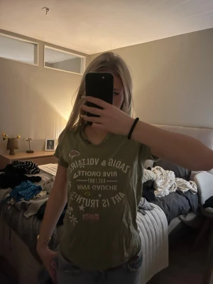 Grön Zadig & Voltaire t-shirt med tryck - Snygg grön t-shirt från Zadig & Voltaire med coolt tryck framtill med blommor, stjärnor, peace-märke och text. T-shirten har rund hals med små knappar och korta ärmar. Perfekt för dig som gillar en avslappnad och trendig stil.