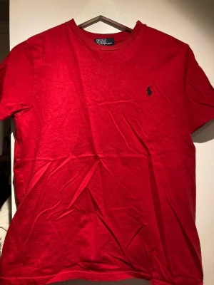 Röd t-shirt från Polo Ralph Lauren - Riktigt snygg röd t-shirt från Polo Ralph Lauren med klassisk broderad logga på bröstet. T-shirten är i mjuk bomull och har en enkel, stilren design med rund hals och korta ärmar. Perfekt för en clean och avslappnad look.