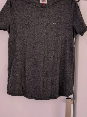 Mörkgrå t-shirt från Tommy Jeans - Snygg mörkgrå t-shirt från Tommy Jeans i storlek L. Klassisk passform med rund hals och diskret logga på bröstet. Tillverkad i mjuk bomull som känns skön mot huden. Perfekt till jeans eller shorts för en clean look.