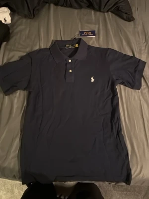 Mörkblå piké från Polo Ralph Lauren - Snygg mörkblå pikétröja från Polo Ralph Lauren med klassisk krage, två vita knappar och den ikoniska broderade loggan på bröstet. Tillverkad i mjuk bomull som känns skön mot huden. Perfekt till jeans eller shorts för en clean look.