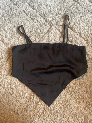 Svart bandana-linnetopp från Divided - Säljer en svart bandana-linnetopp från Divided med smala axelband och smockad rygg. Toppen har en spetsig nederkant framtill och är gjord i ett mjukt, glansigt polyestertyg med lite stretch. Perfekt till sommarens outfits!