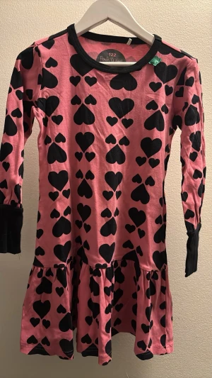 Rosa klänning med svarta hjärtan - Supersöt rosa klänning från Fred's World med svarta hjärtan över hela tyget. Klänningen har lång ärm, svarta muddar och en volang nedtill. Tillverkad i mjuk ekologisk bomull.