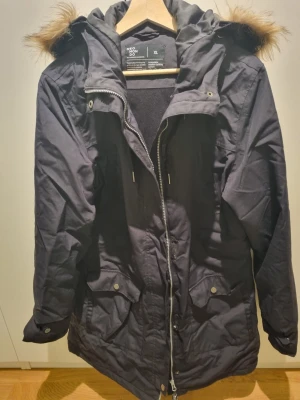 Svart parkasjacka från Neomondo XL - Säljer en svart parkasjacka från Neomondo i storlek XL. Jackan har huva med fuskpäls, dragkedja framtill och praktiska fickor med lock. Perfekt för kalla dagar och har en klassisk, lite längre modell.