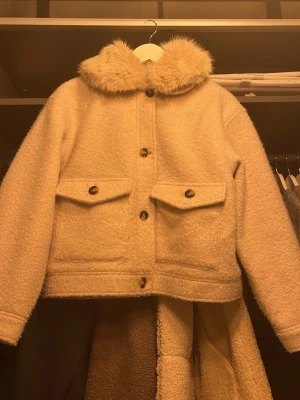 Beige teddyjacka med fuskpäls-krage - Säljer en beige teddyjacka med fluffig fuskpäls-krage och stora fickor framtill. Jackan har knappar hela vägen och en boxig, lite oversized passform. Perfekt för dig som vill ha en mysig och trendig look i höst och vinter.