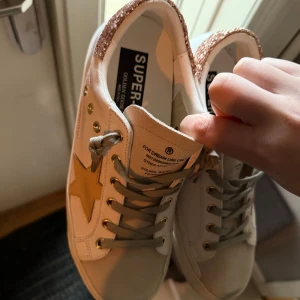 Golden goose sneakers - Storlek 40 men sitter som 39, skorna är använda fåtal gånger så fortsatt mycket bra skick!💗Säljer ett par vita Golden Goose sneakers med beige stjärna på sidan, guldfärgade öljetter och grå snörning. Baktill finns en glittrig detalj i roséguld som ger extra edge. Skorna är i canvas och mocka, med platt sula och rund tå. Pris kan diskuteras 🥰