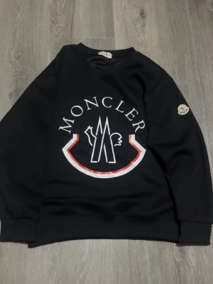 Moncler tröja - Säljer en svart sweatshirt från Moncler med stort vitt och rött logotryck på bröstet och klassisk Moncler-patch på ärmen. Tröjan har rund hals, ribbade muddar och är tillverkad i mjuk bomull. Perfekt för dig som gillar streetwear och vill ha något stilrent men ändå med statement.