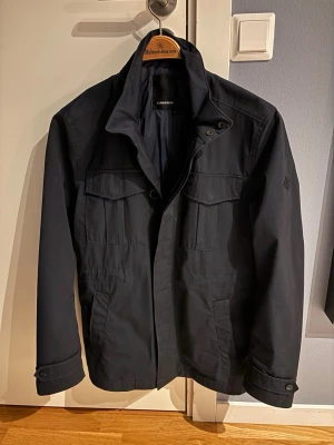 Mörkblå fieldjacket från J.Lindeberg - Säljer en mörkblå fieldjacket från J.Lindeberg med hög krage och fickor framtill. Jackan har knappar och dragkedja. 