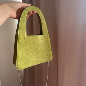 Grön väska med kedja - Snygg grön väska i croco-mönstrad syntet med unik form och silverfärgad kedja som axelrem. Väskan har ett stilrent handtag.