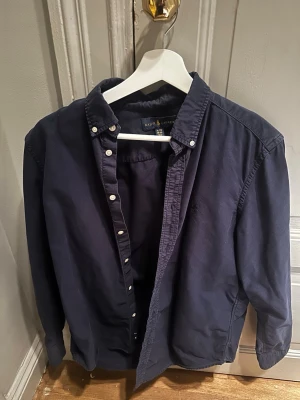 Mörkblå skjorta från Ralph Lauren XL - Klassisk mörkblå skjorta från Ralph Lauren i storlek XL. Skjortan har knappar hela vägen, button-down krage och är långärmad. Tillverkad i mjuk bomull som känns skön mot huden. Perfekt till jeans eller chinos för en clean och stilren look. Passar 178 ish 