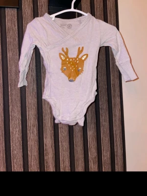 Randig omlottbody med rådjur storlek 62 - Supersöt omlottbody från Lindex med tunna gråvita ränder och ett stort tryck av ett rådjur på magen. Lång ärm och praktisk omlottknäppning. Perfekt för dig som gillar gulliga djurmotiv och mjuka material. Storlek 62