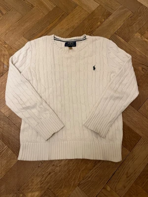 Ralph lauren 10-12 - Vit kabelstickad tröja från Polo Ralph Lauren. Klassisk modell med rund hals och broderad logga på bröstet. Mjuk och skön kvalitet, perfekt till vardag eller mer uppklätt. Använd men i fint skick, inga fläckar eller hål.