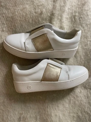 Vita sneakers från Philip Hog med guldresår - Snygga vita sneakers från Philip Hog med bred guldfärgad resår över foten. Skorna har en platt sula. Märkeslogga på sidan av sulan och insidan.