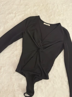 Svart glittrig body  - Säljer en svart body från Glamorous med lång ärm och twistad detalj framtill. Tyget har en glittrig finish som ger en extra snygg touch. Djup v-ringning och åtsittande passform. Perfekt att styla med höga jeans eller kjol.