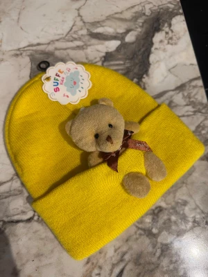 Gul mössa med nalle-detalj Barn - Supersöt gul mössa från Suffe Kids med en mjuk liten nalle fastsydd framtill. Mössan är stickad i syntetmaterial och har en bred uppvikt kant. Perfekt för dig som vill ha något lekfullt och unikt i vinter.