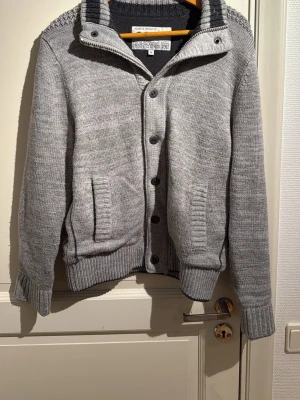 Grå stickad cardigan från Blue-Blooded Rags - Säljer en grå stickad kofta från Blue-Blooded Rags i storlek M. Koftan har ribbade muddar, knappar framtill, fickor och detaljer på axlar och armbågar. Hög krage med kontrastfärg och snygg strukturstickning upptill och på armbågarna.