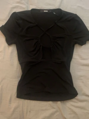 Svart croppad topp från BikBok - Säljer en svart croppad topp från BikBok med korta ärmar och unik cut-out detalj framtill. Toppen har snörning över bröstet och en tight passform som framhäver midjan. Perfekt för dig som vill ha en edgy och trendig look.