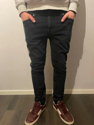 Nudie jeans svarta  - Säljer ett par svarta jeans med raka ben  från Nudie Jeans i storlek W32 L30. Jeansen har klassisk femficksdesign, smal passform och är tillverkade i mjukt bomullstyg med stretch. Snygga detaljer på bakfickorna och diskret logga vid linningen.