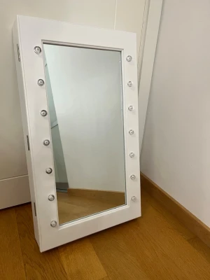 Sminkspegel med LED-belysning och förvaring - Snygg sminkspegel i vitt med inbyggd LED-belysning runt spegeln. Spegeln har ett skåp med svart inredning och flera krokar och hyllor för smink och accessoarer. Perfekt för att organisera dina produkter. Man behöver skruvar för att hänga upp den, och batterier.