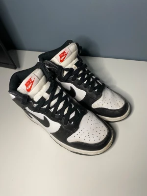 Nike Dunk High svart/vit sneakers - Säljer ett par klassiska Nike Dunk High sneakers i svart och vitt skinn med svarta snören och vit plös med röd Nike-logga. Skorna har perforerad tå, svart swoosh och hög siluett som ger en riktigt clean och tidlös look.