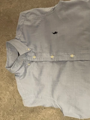 Ljusblå skjorta från Polo Ralph Lauren - Klassisk ljusblå skjorta från Polo Ralph Lauren i slim fit-modell. Skjortan har lång ärm, button-down krage och ikonisk broderad logga på bröstet. Tillverkad i mjuk bomull och stängs med vita knappar framtill. Själv är jag 1,67 och den sitter perfekt på mig men jag skulle säga att den passar runt 160-175 ungefär. Självklart så tvättar jag den innan den fraktas, och priset är ej hugget i sten.