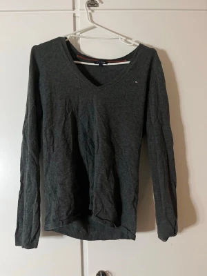 Mörkgrå v-ringad tröja Tommy Hilfiger - Mörkgrå v-ringad tröja från Tommy Hilfiger i storlek L. Klassisk och enkel design med liten logga på bröstet och långa ärmar. Perfekt till höst och vinter, tillverkad i mjukt material som känns skönt mot huden.