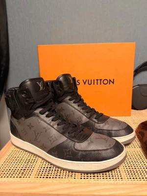 Louis Vuitton monogram high top sneakers - Snygga high top sneakers från Louis Vuitton i svart och grått med klassiskt monogrammönster. Skorna har snörning, vit platt sula och LV-logga på plösen. Tillverkade i exklusivt skinn med detaljer i svart och grått. Perfekta för dig som vill sticka ut med lyxig streetstyle.