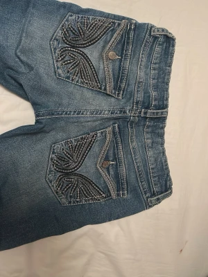 Blå bootcut jeans med broderade fickor - Säljer ett par blå bootcut jeans från Gina Tricot med snygga broderade detaljer på bakfickorna. Jeansen har normal midja, klassisk femficksmodell och dragkedja fram. Materialet är mjukt jeans-tyg som sitter skönt och ger en avslappnad look. Storleken motsvarar S/25W  i vuxen storlek 