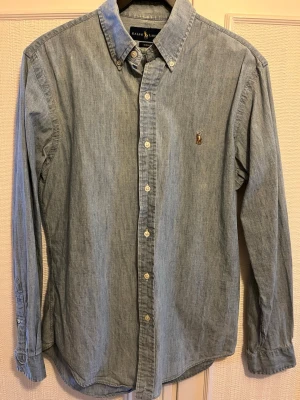 Ljusblå skjorta från Ralph Lauren - Säljer en ljusblå skjorta från Ralph Lauren i slim fit med klassisk button-down krage och broderad logga på bröstet. Skjortan har långa ärmar och vita knappar framtill. Perfekt till jeans eller chinos för en clean look.