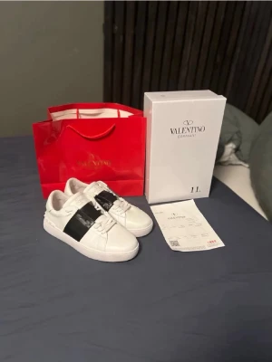 Valentino opens. Mystery gift med på köpet! - Säljer ett par Valentino Garavani Open sneakers i vitt skinn med bred svart läderdetalj över snörningen. Klassisk låg modell med rund tå, vita snören och ikoniska nitar på hälen. Perfekta för dig som vill ha en clean men ändå lyxig look.