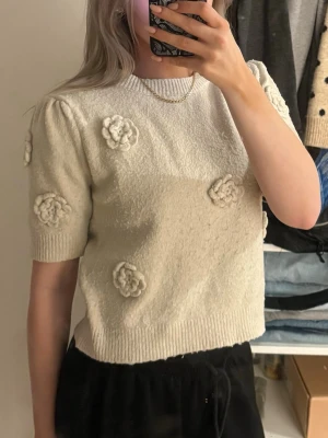Vit stickad topp med blommor från TU - Supersöt vit stickad topp från TU med korta puffärmar och virkade blommor som dekoration framtill och på ärmarna. Tröjan har ribbade muddar vid hals, ärmslut och nederkant. Perfekt för dig som vill ha en gullig och unik look.
