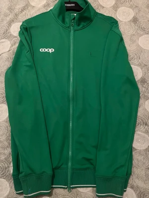 Grön vindjacka från Coop, herr M - Grön vindjacka från Coop i herrmodell, storlek M. Jackan har hel dragkedja framtill, ribbade muddar med vit rand och hög krage. Coop-logga i vitt på bröstet och ryggen. Tillverkad i polyester för en sportig look.