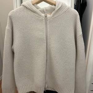 Vit ribbad stickad zip tröja - Mysig vit stickad zip tröja med ribbad struktur och dragkedja framtill. Tröjan har huva och långa ärmar, perfekt för kyliga dagar. Passar dig som gillar en enkel men ändå stilren look.