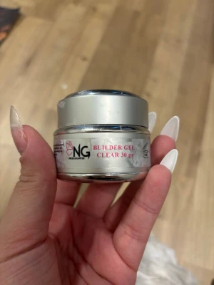 NG Builder Gel Clear 30g - NG Builder Gel Clear är ett genomskinligt bygggele för naglar i en rund burk på 30 gram. Perfekt för att skapa starka och snygga nagelförlängningar. Burken är silverfärgad med vit etikett och rosa text. Gelen är klar och har en tjock konsistens som är lätt att arbeta med. Knappt använd 