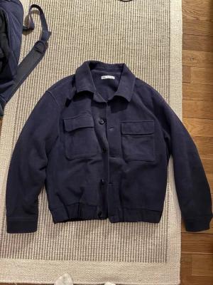 Mörkblå overshirt från Zara - Säljer en mörkblå overshirt/ jacka från Zara med två stora bröstfickor och knäppning framtill. Jackan har klassisk krage och är tillverkad i mjukt bomullsmaterial. Skulle säga att den är mer vårjacka än overshirt. Storlek L men sitter som M
