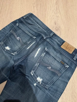 Selvedge Nudie Jeans - Säljer ett par sjukt feta selvedge jeans från Nudie Jeans med slitna detaljer och kontrastsömmar. Jeansen har fem fickor, normal passform och raka ben. Materialet är slitstarkt denim i bomull och färgen är snyggt tvättad blå med ljusare partier.