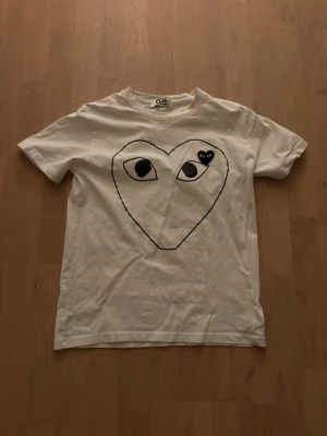 Vit Comme des Garçons Play t-shirt - Säljer en vit t-shirt sopm har det ikoniska hjärtmotivet med ögon från Comme des Garçons Play på bröstet. Klassisk passform med rund hals och korta ärmar. Tillverkad i mjuk bomull som är skön att bära. Perfekt för dig som gillar streetwear och vill sticka ut. Storlek M men sitter som S. Tröjan är använd 5 gånger och är som ny, inga deffekter eller kosmetiska skador.