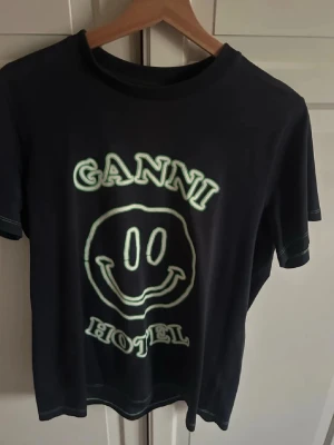 Svart Ganni t-shirt med smiley - Svart t-shirt från Ganni med stort smiley-tryck och texten 'Ganni Hotel' i vitt och grönt på framsidan. Klassisk rund hals och korta ärmar. Tillverkad i mjuk bomull som känns skön mot huden. Perfekt för en chill och trendig look.