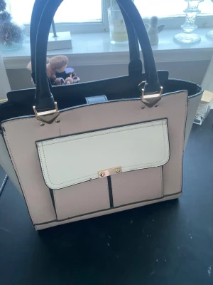 Ljusrosa och svart handväska med detaljer - Snygg handväska i ljusrosa och svart med vita detaljer och guldfärgade metalldelar. Väskan har två rejäla handtag, en stor ytterficka med lock och tryckknapp samt rymligt huvudfack. Stilren och modern look. (Lite fläckig men inget man tänker på, se bild 3)