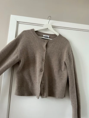 Beige merinoullskofta från & Other Stories - Säljer en beige kofta från & Other Stories i 100% merinoull. Koftan har rund hals, ribbade muddar och knäppning med matchande knappar framtill. 