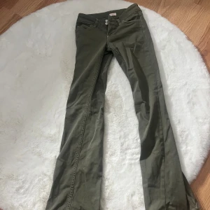 Olivgröna bootcut byxor - Säljer ett par olivgröna byxor med bootcut-snitt. Byxorna har låg midja och klassisk femficksdesign. Tillverkade i ett mjukt bomullsmaterial som ger en skön känsla och snygg passform. Perfekta för dig som gillar en avslappnad men trendig look.