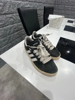 Svarta Adidas Gazelle sneakers - Säljer ett par klassiska Adidas Gazelle sneakers i svart mocka med vita detaljer och de ikoniska tre ränderna på sidan. Skorna har vit sula, breda vita skosnören och en låg profil. Perfekta för dig som gillar retrostil och streetwear.