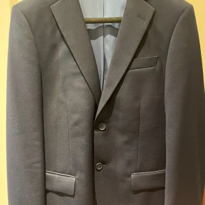 Mörkblå kostym från Dressmann, strl 54 - Säljer en stilren mörkblå kostym från Dressmann. Kavajen har två knappar, klassiska fickor och smal passform. Byxorna är raka med bälteshällor och matchar perfekt till kavajen.