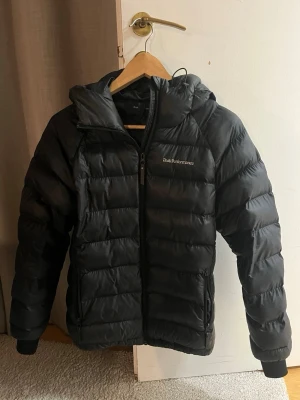 Svart pufferjacka från Peak Performance - Säljer en svart pufferjacka från Peak Performance med huva och dragkedja framtill. Jackan har en quiltad design, två sidofickor med dragkedja och ribbade muddar vid ärmsluten. Perfekt för kalla dagar och riktigt snygg till vardagsoutfiten.