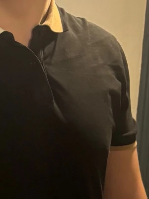 Svart kortärmad t-shirt från BOSS - Snygg svart t-shirt från BOSS med korta ärmar och kontrasterande beige detaljer på krage och ärmslut. Kragen har en cool logga med texten 'BOSS' i vitt och svart. Perfekt för en clean och stilren look.