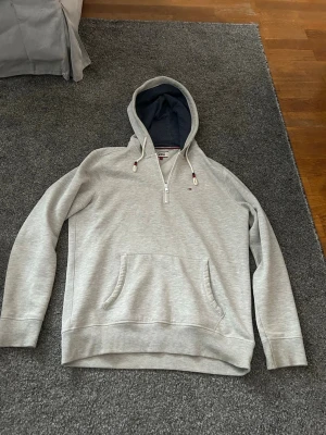 Tommy Jeans Half-Zip Hoodie – Y2K / Vintage - Grå hoodie från Tommy Jeans (Tommy Hilfiger) med halv dragkedja. Tröjan har huva med mörkblått foder, känguruficka framtill samt klassisk Tommy-flagga på bröstet. Tillverkad i mjukt och följsamt sweatshirttyg med ribbade muddar i ärmslut och nederkant.  Modellen är från sent 1990-tal / tidigt 2000-tal (Y2K-era) och är ett originalplagg från Tommy Jeans-linjen. Tidlös design som fungerar både till vardags och som del av en streetwear- eller Y2K-inspirerad outfit.  Plagget är i mycket bra skick!