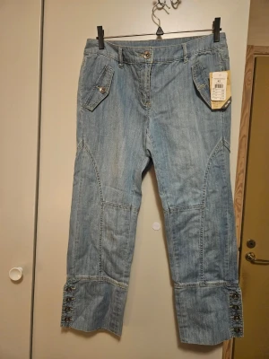 Blå jeansbyxor med knappar, Casual fit, strl 40 - Snygga ljusblå jeansbyxor från Kappahl med normal midja och Casual passform. Byxorna har dekorativa knappar vid bensluten och coola fickor framtill med knappdetaljer. Tillverkade i stretchigt jeansmaterial för extra komfort.