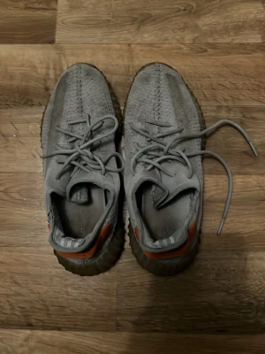 Adidas Yeezy Boost 350 V2 Grå/Orange - Säljer ett par Adidas Yeezy Boost 350 V2 i grått med orange detaljer på hälen och sidan. Skorna har stickad ovandel i syntet, snörning och en chunky, genomskinlig sula. Perfekta för dig som gillar streetwear och vill sticka ut med en unik färgkombo.
