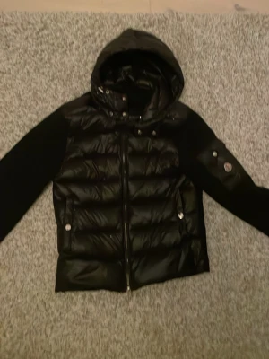 Svart cardigan från Moncler - Säljer en svart cardigan från Moncler med huva och dragkedja framtill. Jackan har stickade ärmar och en ficka med Moncler-logga på ena armen. Materialmix med blank polyester och stickad struktur på ärmarna. Perfekt för kyliga dagar.