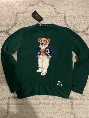 Grön stickad tröja Polo Ralph Lauren - Mörkgrön stickad tröja från Polo Ralph Lauren med ikonisk björn på bröstet. Tröjan har rund hals, ribbade muddar och RL-broderi nedtill. Perfekt för dig som vill ha en klassisk men lekfull look.